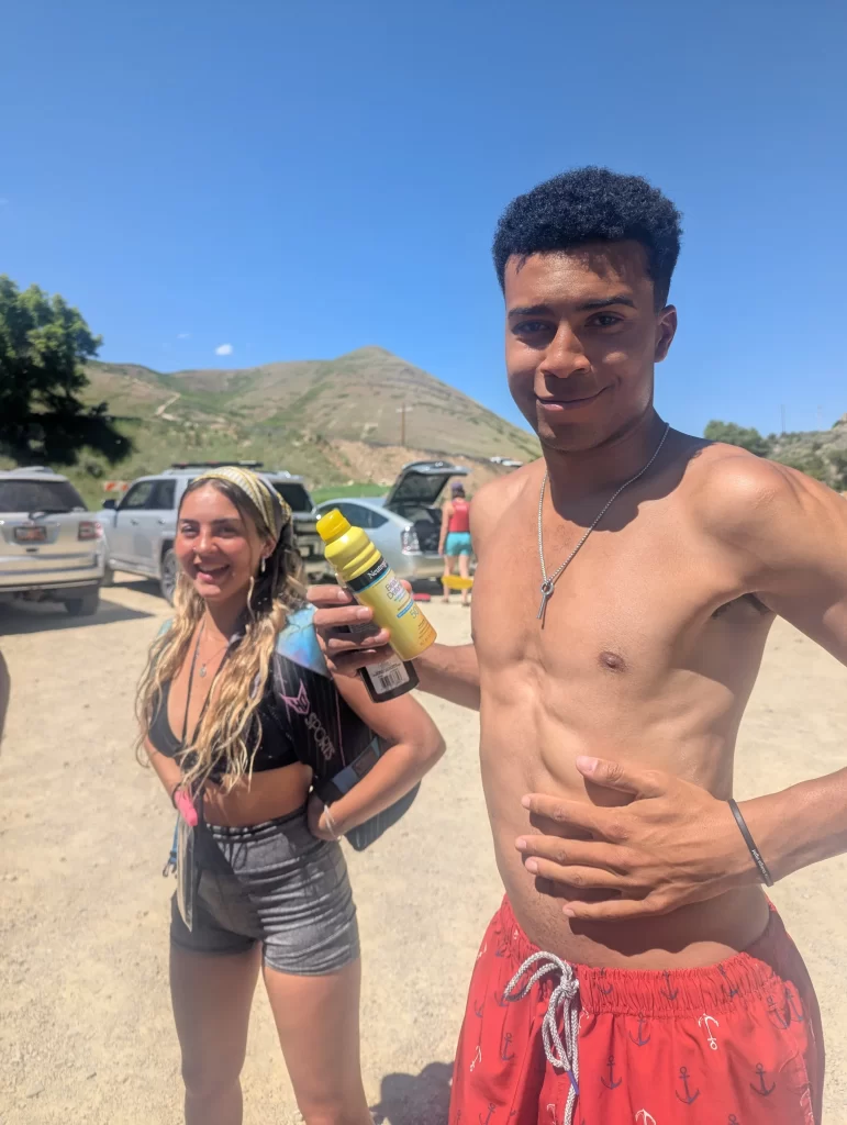 Two young adults standing outdoors near their cars, preparing for a tubing adventure on the Provo River. One is holding a bottle of sunscreen, dressed in swimwear, while the other is wearing shorts and a swimsuit top, set against a blue sky and scenic mountains.