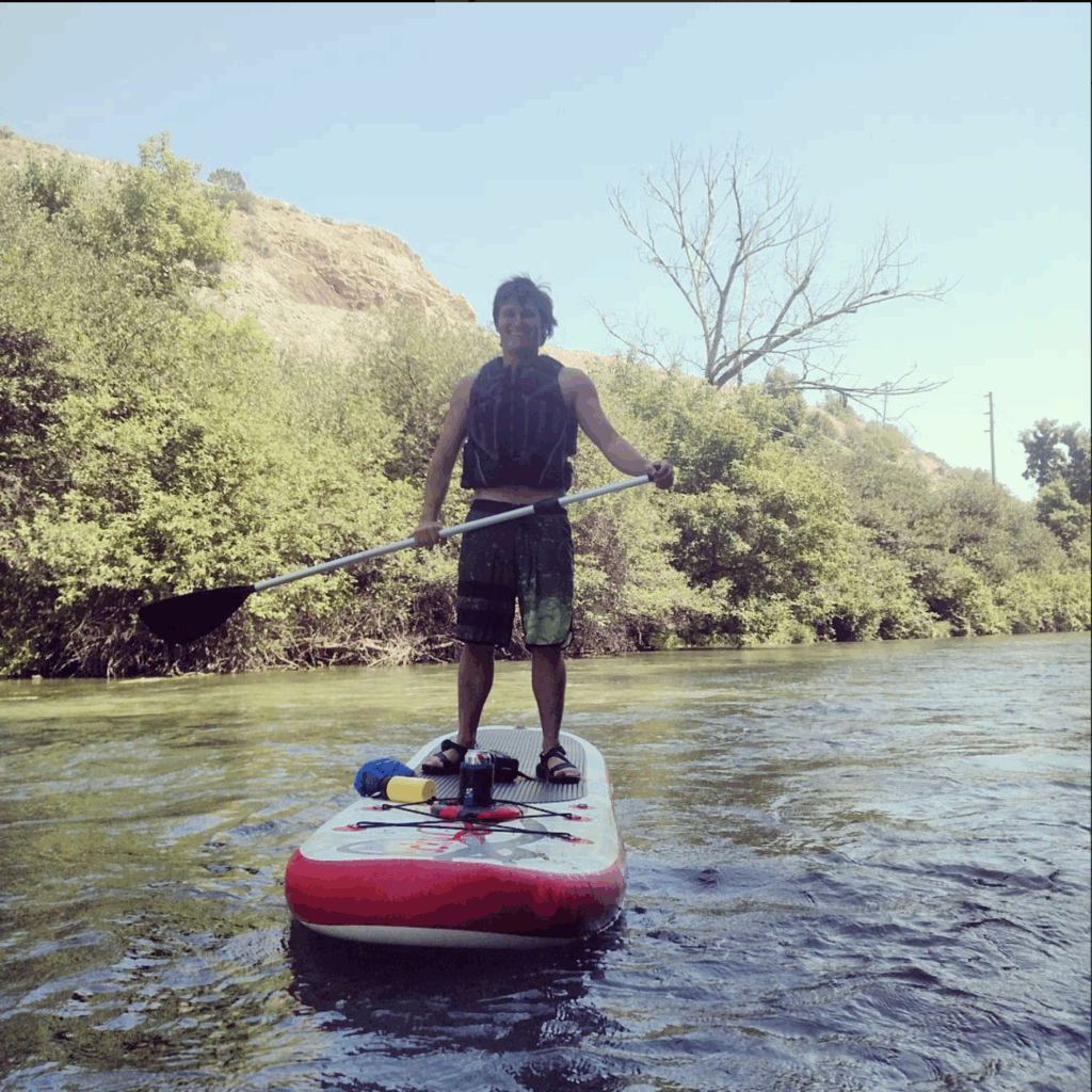 A person standing on a paddle board in a river surrounded by greenery, holding a paddle and wearing a life jacket.